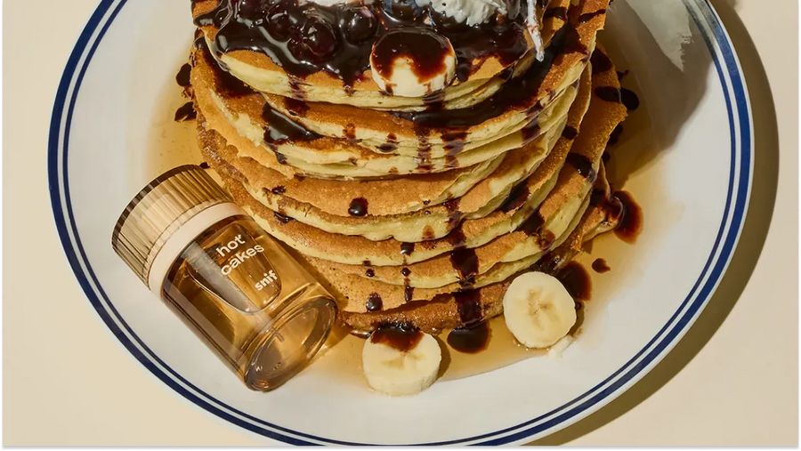 Pancake with honey and banana slices on a plate beside Snif Hot Cakes perfume, symbolizing fragrance sweetness and how long perfume lasts on skin.